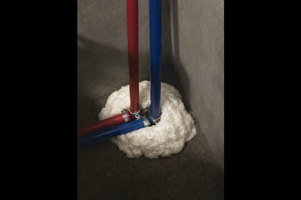 Red and blue RV PEX water lines passing through a floor opening sealed with expanding foam insulation to prevent mice from entering an RV.